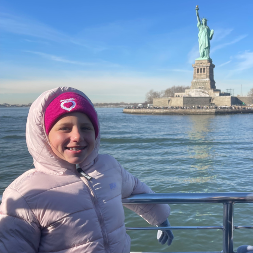 A child in a pink coat and magenta DonateLife beanie stands on a boat in front of the Statue of Liberty. The sky is clear and blue.