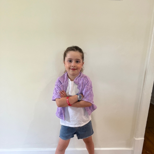 young girl in a white shirt and a purple short sleeved jacket. She's standing with her feet wide apart, arms crossed, smiling at the camera.