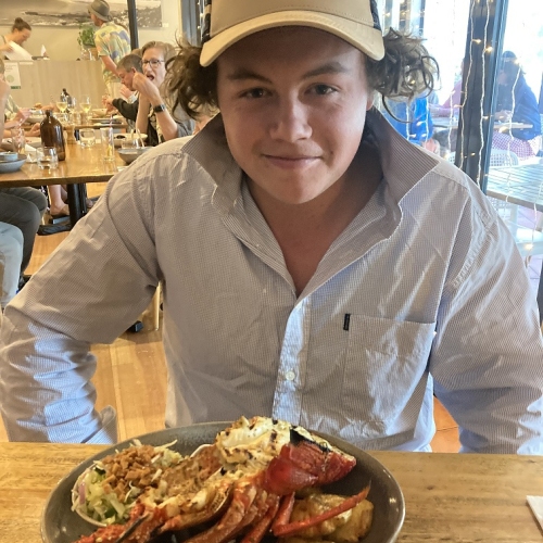 20 year old Harvey wearing a tan and black cap with a blue long sleeve collared shirt on, smiling in front of a plate of lobster