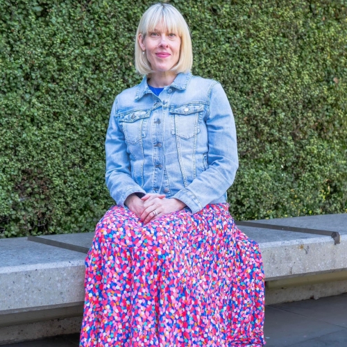 Photo of Jessica Chappel who had a liver transplant. Jessica is wearing a denim jacket with a pink foral skirt, sitting on a concrete bench in front of a green hedge