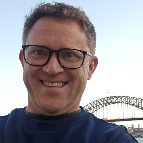 A man in his forties wearing glasses and a blue shirt smiles while posing in front of the Sydney Harbour Bridge. 