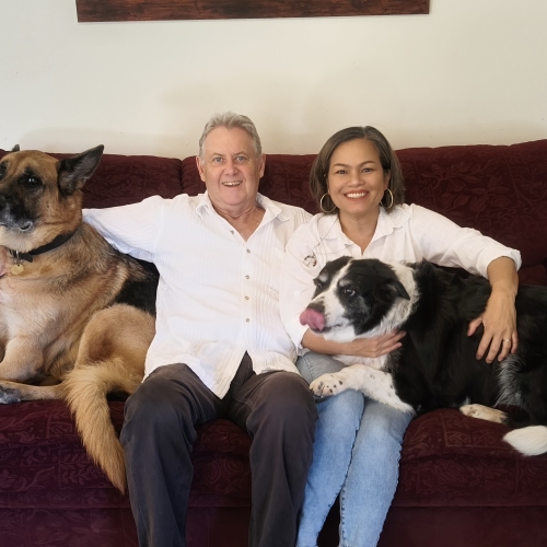A man and a woman sit on a maroon couch, smiling at the camera while a black and white dog, licking its nose, rests in her lap.