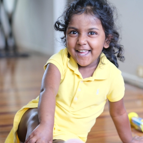 Little girl sitting inside on a wooden floor. She is smiling at the camera and is wearing a yellow dress.