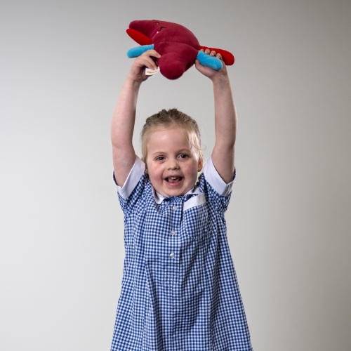 A young girl in a blue and white striped school uniform smiles broadly and holds a stuffed toy in the shape of a liver in the air above her head.