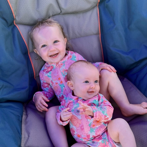 A toddler and a baby in pink swimmers sitting together in a camping chair