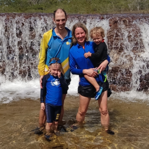 A man and woman pose with their two young boys in front of a large waterfall on a sunny day. The family is smiling and wearing swimmers.