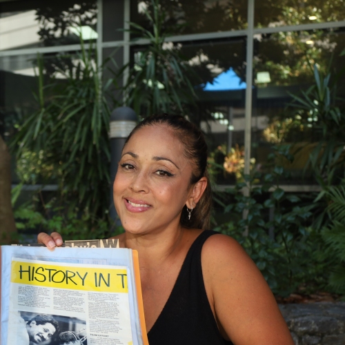 A dark-skinned woman in her 40s sits at a table. She is holding up a folder with newspaper clippings against a backdrop of greenery.  