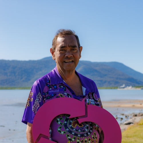 A person in a colourful shirt holds a large pink DonateLife heart symbol against a serene lakeside backdrop with mountains.