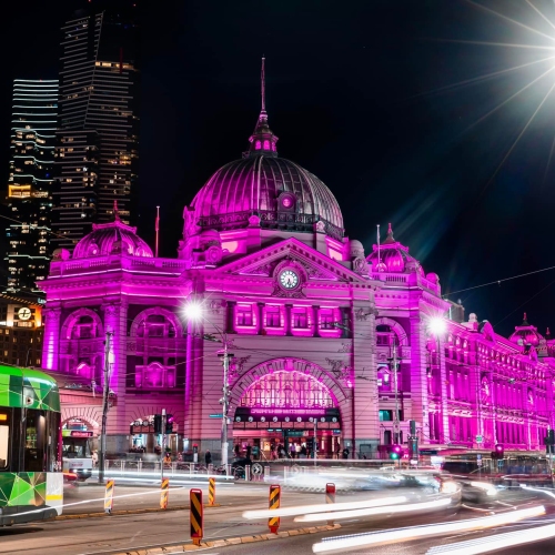 Flinders Street Station in Melbourne lit up in magenta at night against a city backdrop. A tram and care traffic are in the foreground.