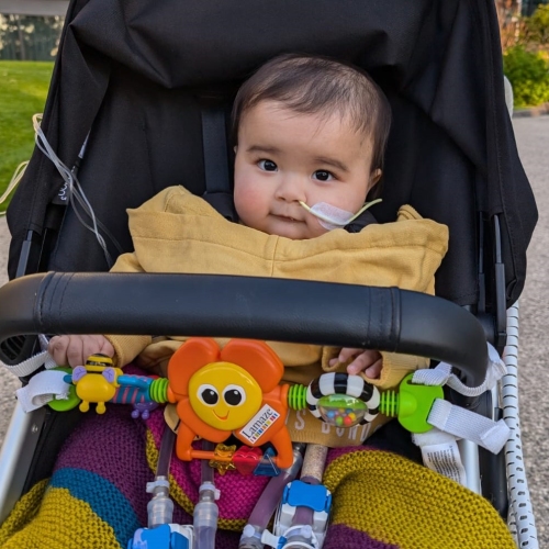 A baby boy wearing a nasal cannula sits in a pram covered by a mustard coloured blanket. There are toys attached to the pram handle and in the background is a park setting.