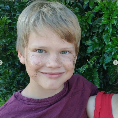 A young boy with blonde hair and blue/green eyes smiles at the camera against a backdrop of greenery.