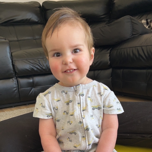  A young child wearing a light-coloured onesie with small vehicle prints and white socks is sitting on a colourful foam mat. The child is holding a rainbow-colored pop-it toy. The background features black leather couches.
