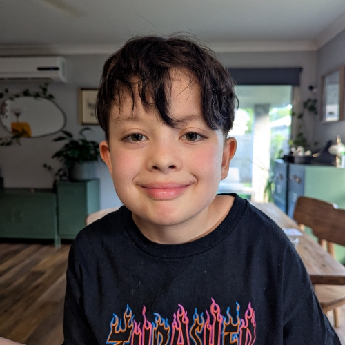 Child in a black “Thrasher Magazine” t-shirt stands indoors. Background features wooden floors, green furniture, and plants.