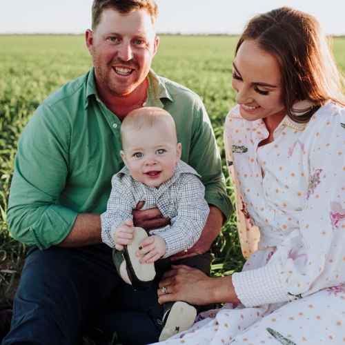 A family of 3 sitting in a grassy field. The adult on the left wears a green shirt and holds a baby in a checkered shirt and light-coloured pants. The adult on the right wears a white dress with floral patterns.