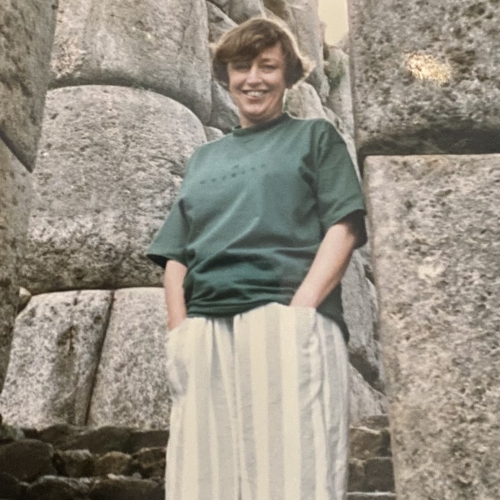 A person standing on stone steps between large, ancient stone walls made of massive, irregularly shaped stones. The individual wears a green shirt and white pants with vertical stripes.