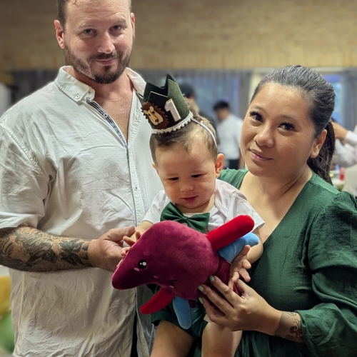 3 people posing indoors at a celebration. A tattooed person in a white shirt stands on the left. In the centre, a child wearing a green crown with the number '1' holds a red stuffed toy in the shape of a liver. On the right, a person in a green dress holds the child. Balloons are visible in the background.