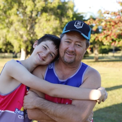 A young person and an older person, both wearing sleeveless sports jerseys, stand together in a park. The young person smiles with their head resting on the older person's shoulder, arms wrapped around them. The older person, wearing a cap, smiles at the camera. Trees, grass, and other people are visible in the background.