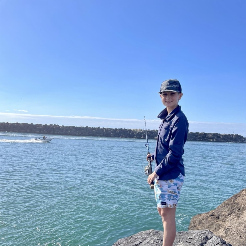 A child standing on a rocky shoreline holding a fishing rod, wearing a dark long-sleeve shirt, patterned shorts, flip-flops, and a cap. Calm blue water and a boat are visible in the background, with a tree-lined shore under a clear blue sky."