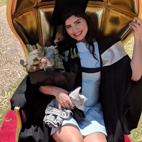 Person in graduation cap and gown seated on a large ornate golden chair, holding a bouquet of flowers wrapped in black paper, smiling at the camera with grass and people visible in the background. There is white text on a pink background that reads ‘Scarlett became an organ donor’.