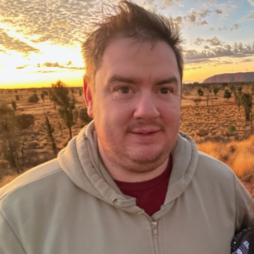 A person stands in a rugged landscape during sunset, with Uluru in the distance and vibrant clouds overhead.