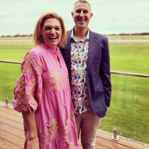 Two people standing on a wooden deck with a glass railing, overlooking a large grassy field under a cloudy sky. One person is wearing a bright pink dress with gold embroidery, and the other is dressed in a patterned shirt, light-coloured pants, and a dark blazer.