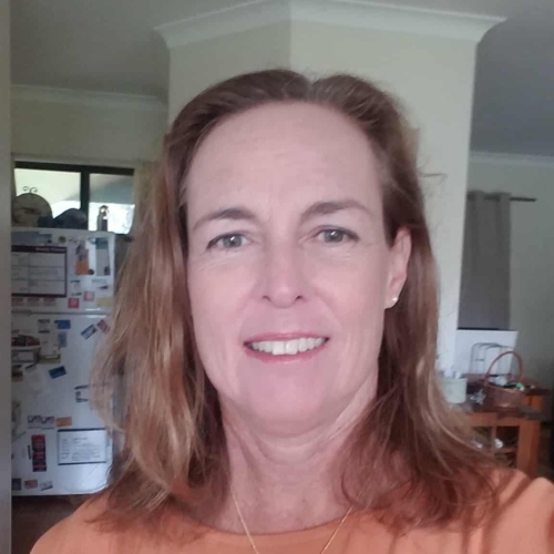 A person wearing an orange top with a round gold pendant necklace, standing indoors. In the background, there is a refrigerator covered with magnets and papers, and a dining table with various items on it.