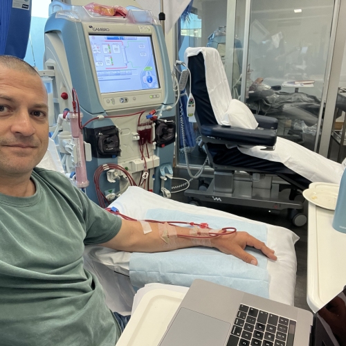 A person is seated in a medical facility, undergoing dialysis treatment. The individual has an IV line inserted into the arm, connected to a dialysis machine with various tubes and a digital display. The person is using a laptop placed on a tray table in front of them. There is also a water bottle and some personal items on the table. In the background, there are other medical chairs and equipment visible through glass partitions. 