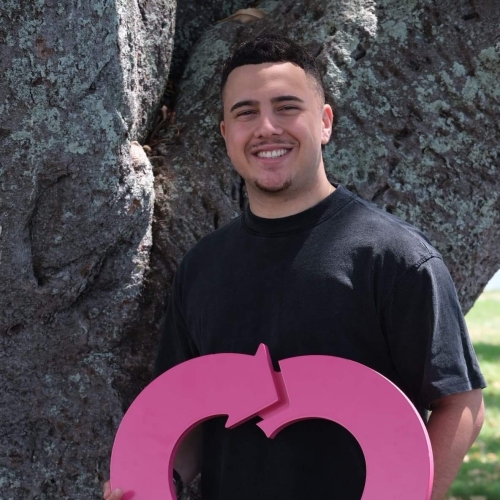 A man in his 20s wearing a black shirt poses in front of a tree. He is smiling and holding a DonateLife wooden logo. 