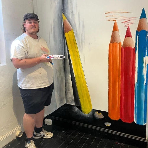 Person painting a colourful wall mural of oversized pencils, including a large yellow pencil and 3 upright colored pencils on a white wall.