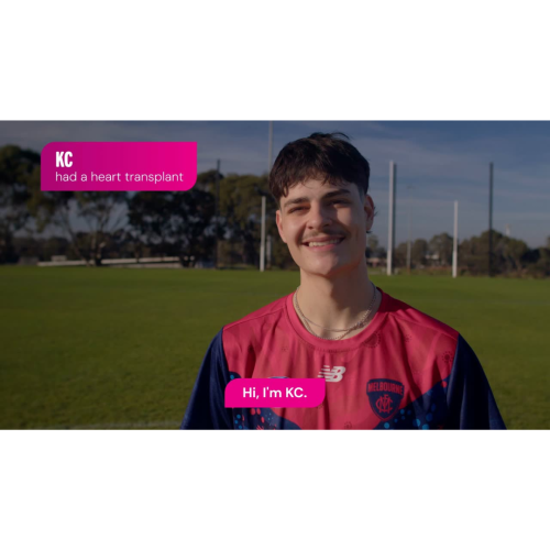 A person stands on a grassy sports field with tall goal posts and trees in the background under a clear sky, wearing a red and navy AFL jersey with a Melbourne club emblem and layered chain necklaces, with short hair styled in a mullet and a moustache, facing the camera. Bright pink text overlays appear, with one in the upper left reading “KC had a heart transplant” and another centered near the bottom reading “Hi, I’m KC.”