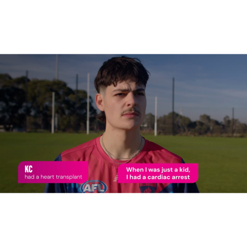 A person stands outdoors on a grassy sports field with tall goal posts in the background under a clear sky, wearing a red and navy sports jersey with an AFL logo and a Melbourne club emblem along with chain necklaces, medium-length hair in a mullet-style cut and a moustache. At the bottom of the image, two bright pink text boxes read: “KC had a heart transplant” on the left and “When I was just a kid, I had a cardiac arrest” on the right.