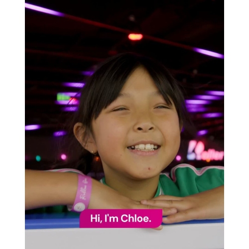 A child leans on a railing in a brightly lit indoor venue with colourful lights in the background, wearing a wristband and smartwatch, while on-screen text reads “Chloe had a liver transplant” and “Hi, I’m Chloe.”