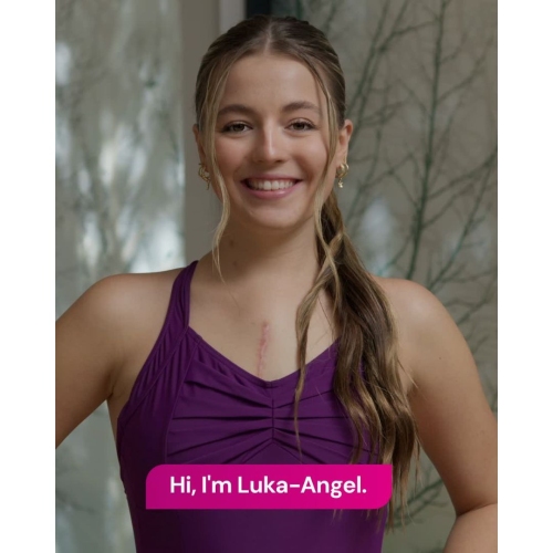 Close-up portrait of a person in a purple sleeveless top with hair tied back, standing indoors. A vertical chest scar is visible, and a pink banner reads: “Hi, I’m Luka-Angel.”
