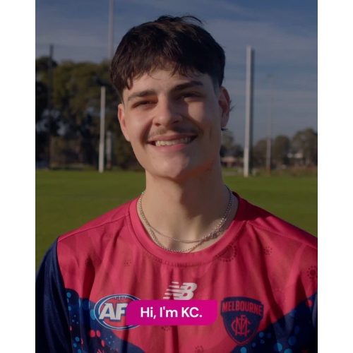 A young person wearing a red Melbourne Football Club jersey stands outdoors on a sports field, with text on screen reading “Hi, I’m KC.”