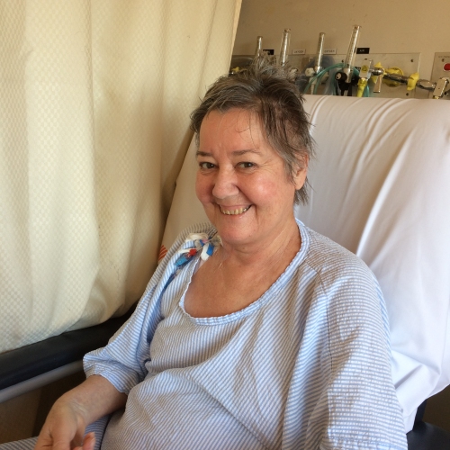 A person is smiling while sitting in a hospital bed wearing a light blue hospital gown. They are holding a pen and paper, possibly writing or drawing. Medical supplies and equipment are visible in the background on a shelf, and a curtain partially covers the left side of the image.