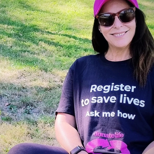 Person sitting on grass outdoors wearing a pink DonateLife cap, sunglasses and a black T-shirt displaying the message “Register to save lives. Ask me how."