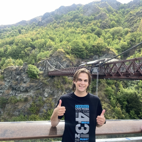 Person standing at a viewing platform giving two thumbs up, with the Kawarau Bungy Bridge spanning a rocky gorge and forested mountains in the background.