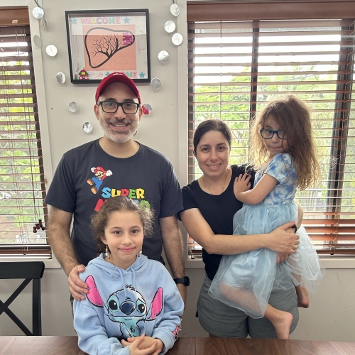 A family of four poses indoors beside a dining table, with one child seated in front and another being held, while a framed artwork hangs on the wall behind them.