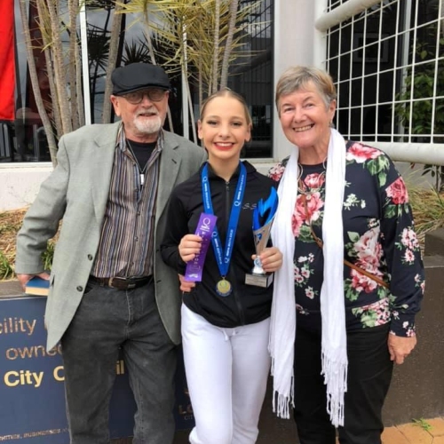 A young athlete stands outdoors holding a trophy, ribbon and medal, posing with two older adults in front of a building and landscaped plants.
